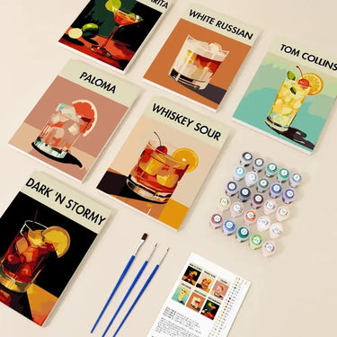 Classic Cocktails -numeromaalaussetti, jossa kuusi 20x15 cm drinkkiteemaista maalausta nimillä, maalipurkit, pensselit ja ohjeet pöydälle aseteltuna.